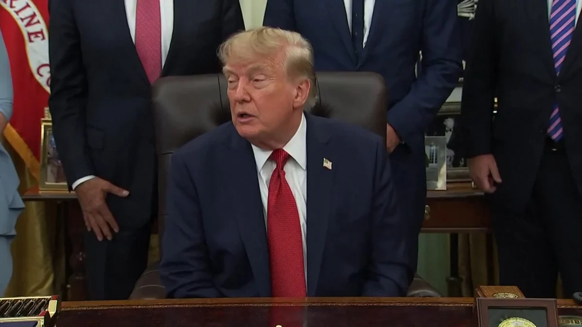 Donald Trump seated at desk with officials behind him