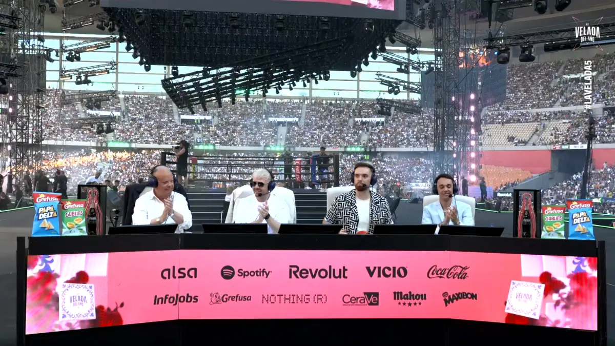 Commentators at sports event, large audience in background.