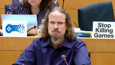 Man speaking at hearing with Stop Killing Games sign
