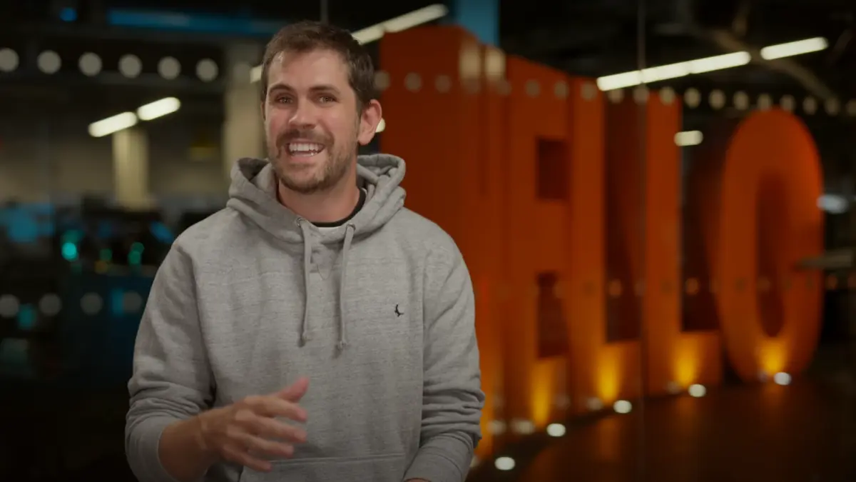 Smiling person in gray hoodie near orange 'HELLO' sign.