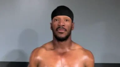 Shirtless man wearing black durag against gray wall