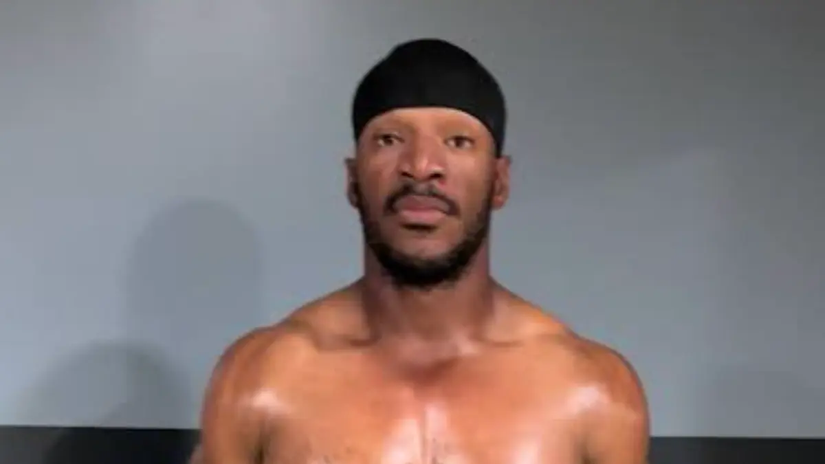 Shirtless man wearing black durag against gray wall
