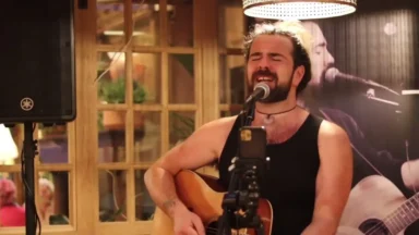 Bearded musician sings while playing acoustic guitar indoors.