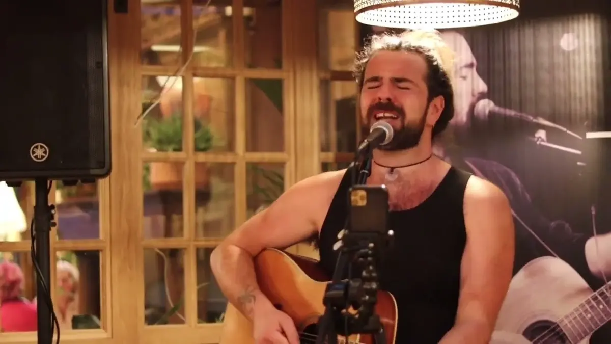 Bearded musician sings while playing acoustic guitar indoors.